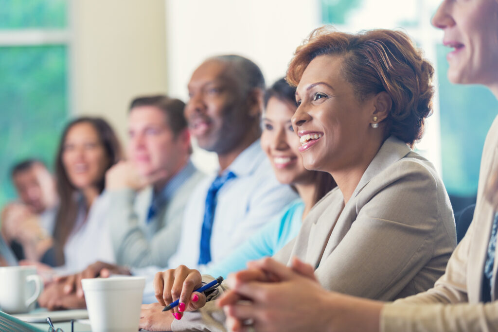 Mid adult African American businesswoman is smiling while attending business conference. She is listening to speaker in seminar while she sits in a row with other diverse professional businesspeople. Woman is wearing business casual clothing and is taking notes. She's drinking a cup of coffee.